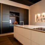 Wandkast in een keuken uitgevoerd in donkergrijs decoratief plaatmateriaal met een gemêleerd decor met de structuur van luxe zijdedraad, de FB08 Reflex van Cleaf. De fronten van het kookeiland zijn uitgevoerd in mat wit plaatmateriaal HM00 Piombo van Cleaf en het werkblad in solid surface decor Weathered Wood van Durasein.